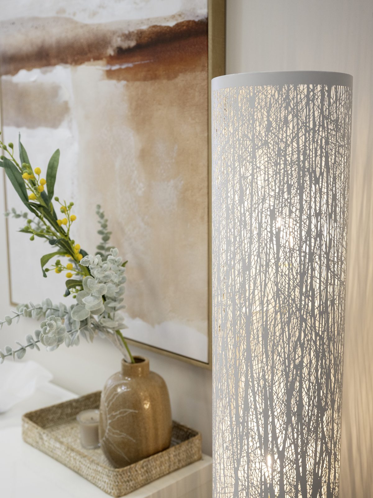 A textured floor lamp beside a vase of eucalyptus in front of a soft abstract painting