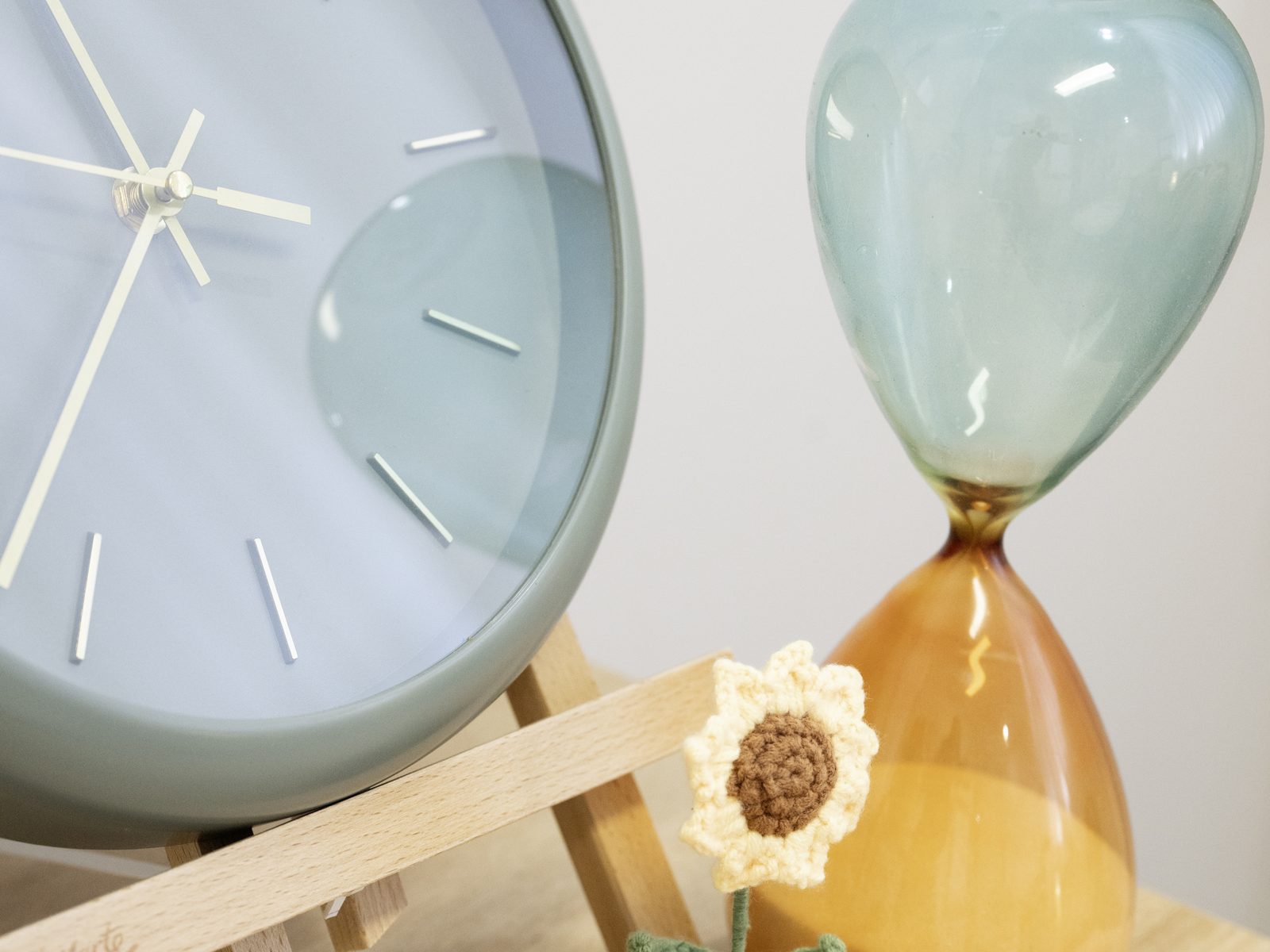 A sage green clock, glass hourglass and crocheted sunflower on a shelf