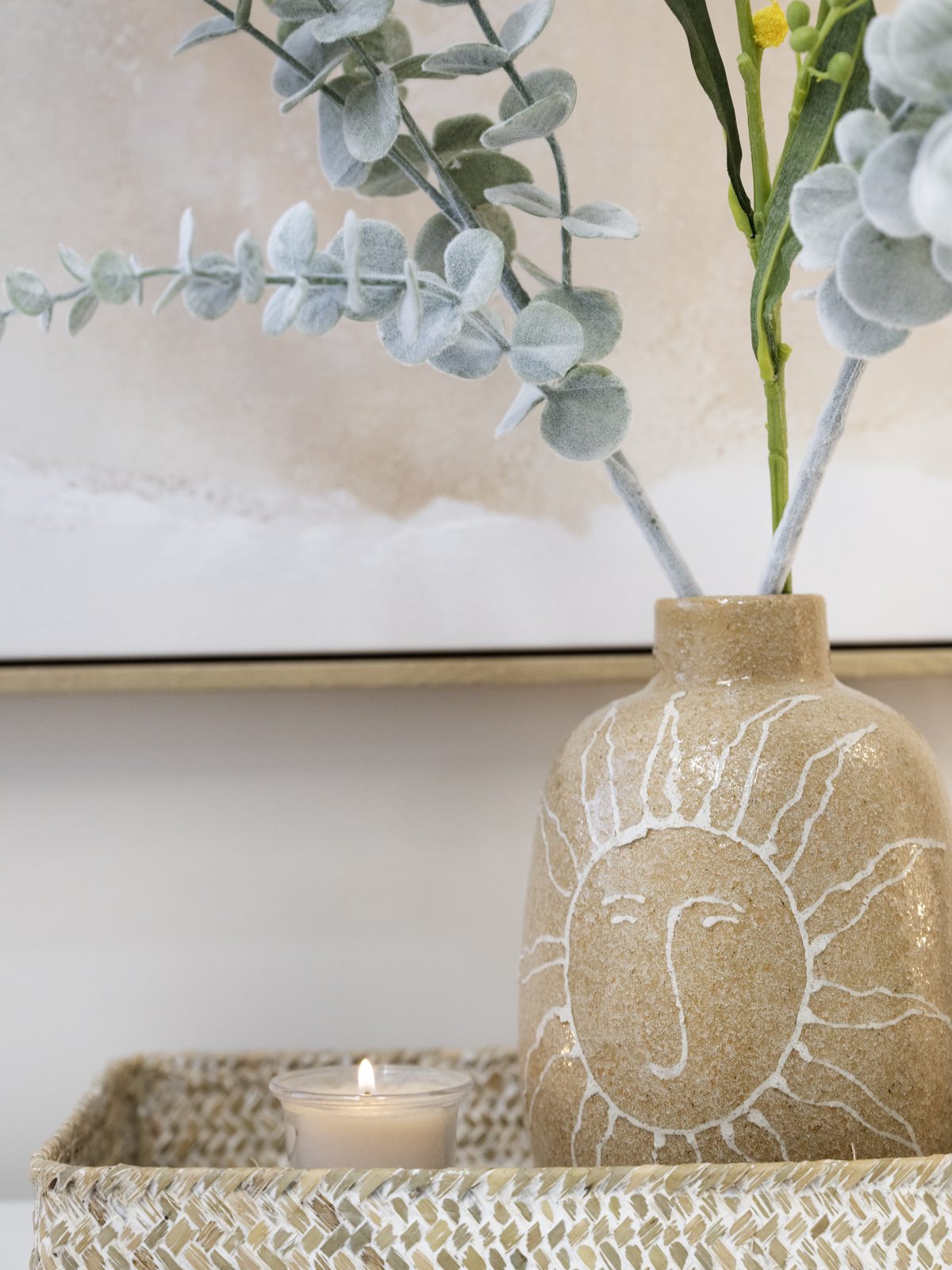 A hand-drawn sun vase holding eucalyptus and wattle on a woven tray with a tealight